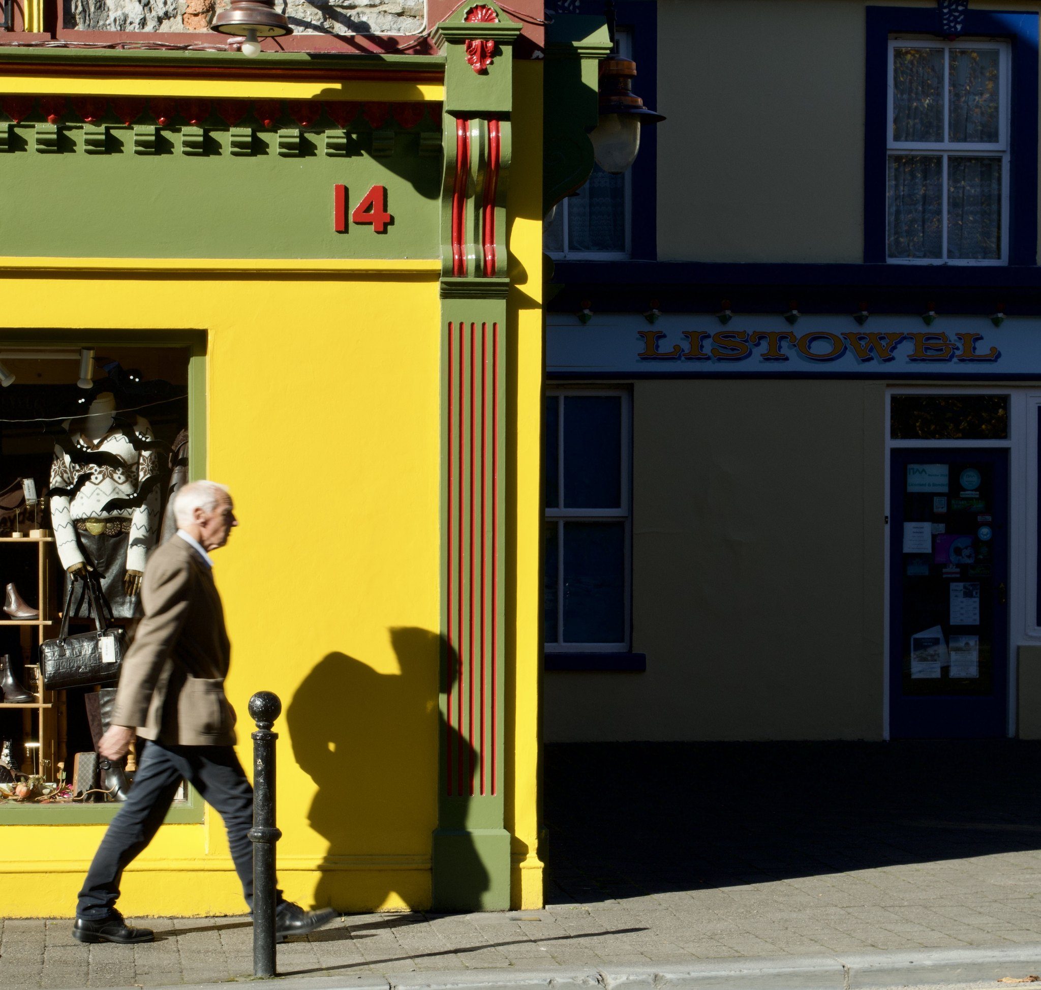 Colour photograph of an older man in a tweed jacket walking past a bright yellow shopfront, number 14, his shadow on the wall beside him, in Listowel, County Kerry
