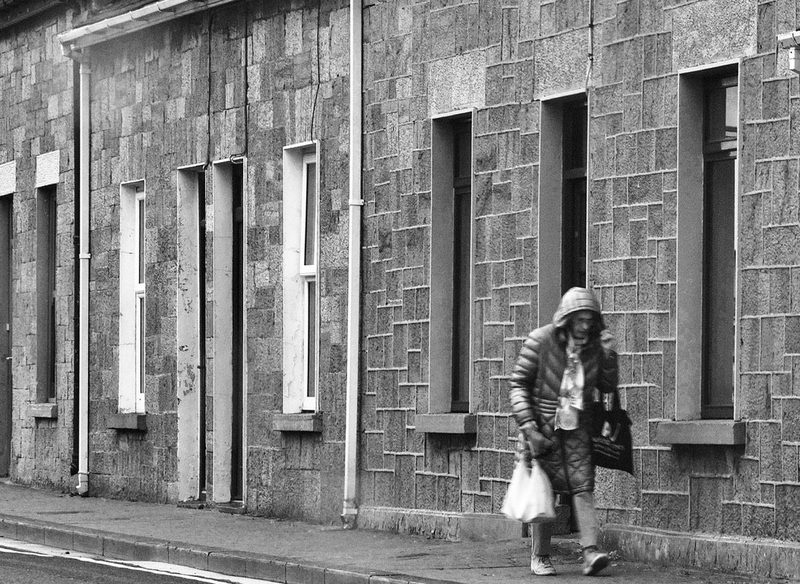 Black and white photograph of a woman in a hooded coat carrying her shopping past the stone fronts of terraced houses on Market Street, Listowel, County Kerry