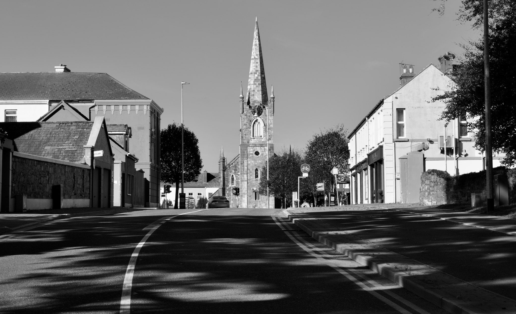 Black and white photograph of an empty street in low morning sun, tree shadows across the road leading to the Gothic spire of St John's, Listowel, County Kerry
