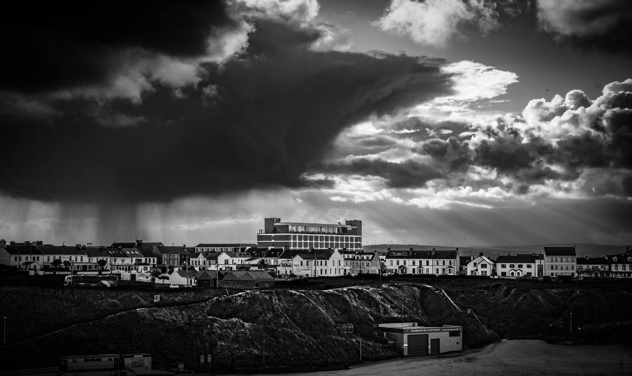 Black and white photograph of a dark storm cloud and a shaft of rain over the Atlantic, with sunlight breaking over the seafront terraces of Ballybunion, County Kerry